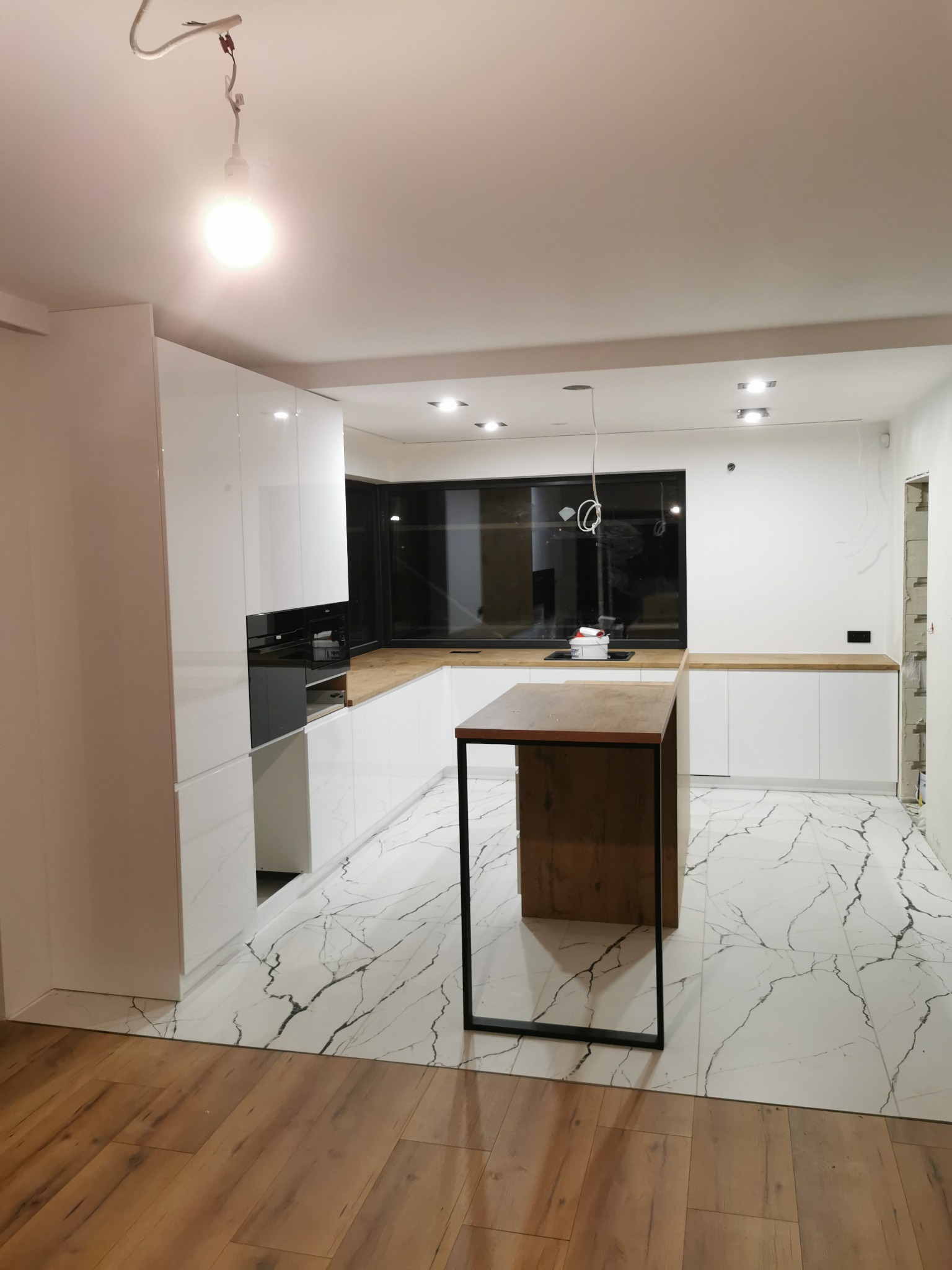 Nowoczesna biała kuchnia z drewnianym blatem i wyspą na tle marmurowej podłogi, widoczne niedokończone prace wykończeniowe i surowa żarówka.