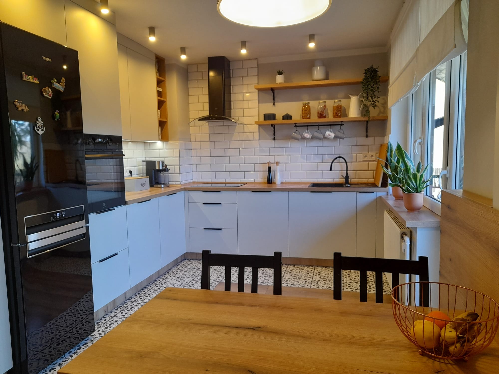 Jasna kuchnia z białymi szafkami i drewnianym blatem. Na ścianie białe płytki, drewniane półki z dekoracjami. Na pierwszym planie stół z owocami.