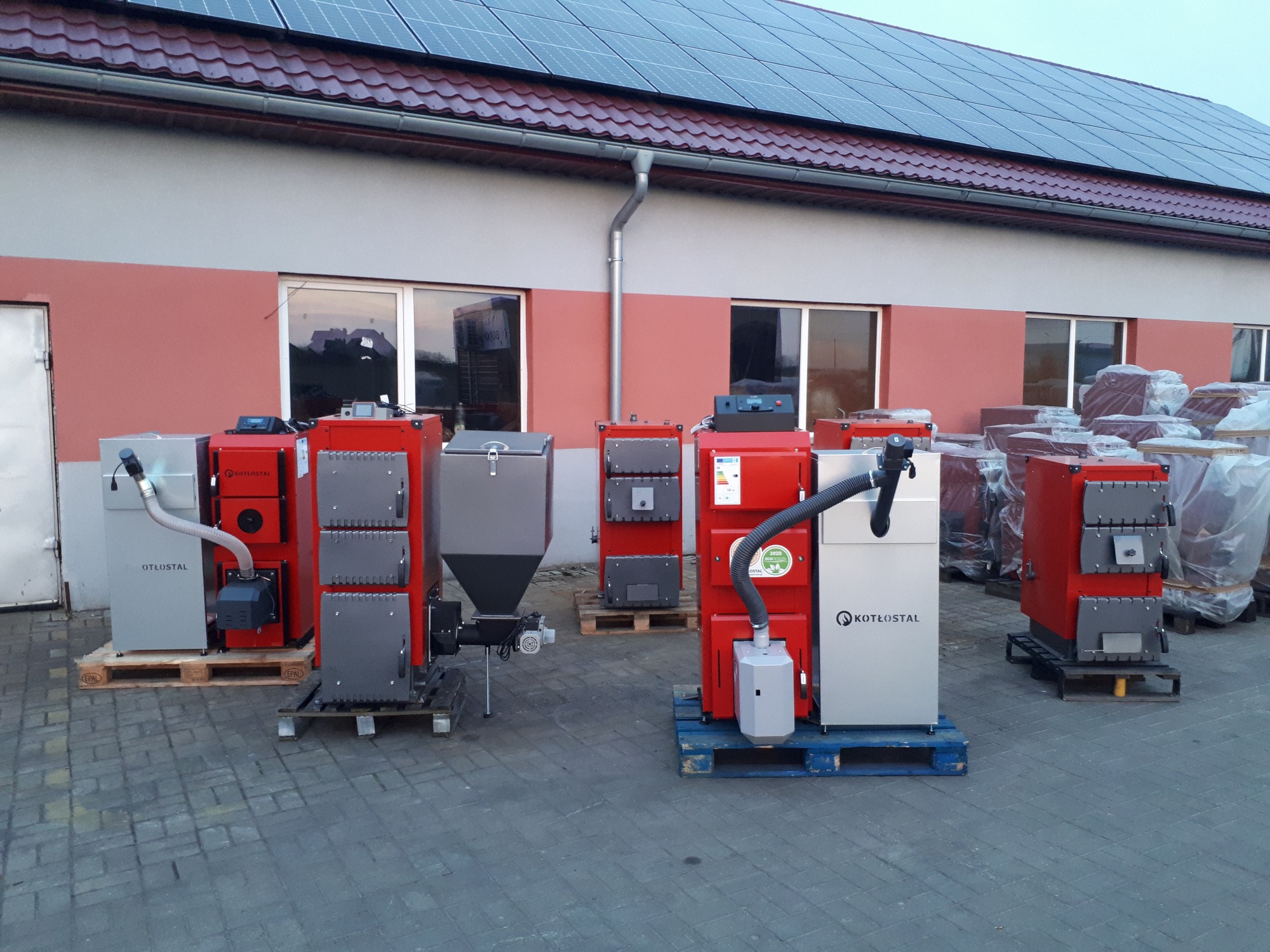 Różne modele kotłów na paliwo stałe marki Kotłostal i Otłostal, ustawione na paletach przed budynkiem z panelami słonecznymi na dachu.