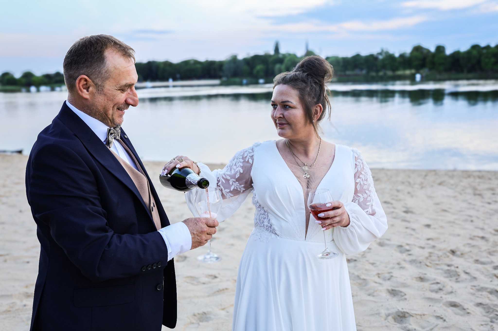 Elegancka para na plaży wznosi toast szampanem; kobieta w białej sukni, mężczyzna w garniturze. Romantyczna sceneria z jeziorem w tle.