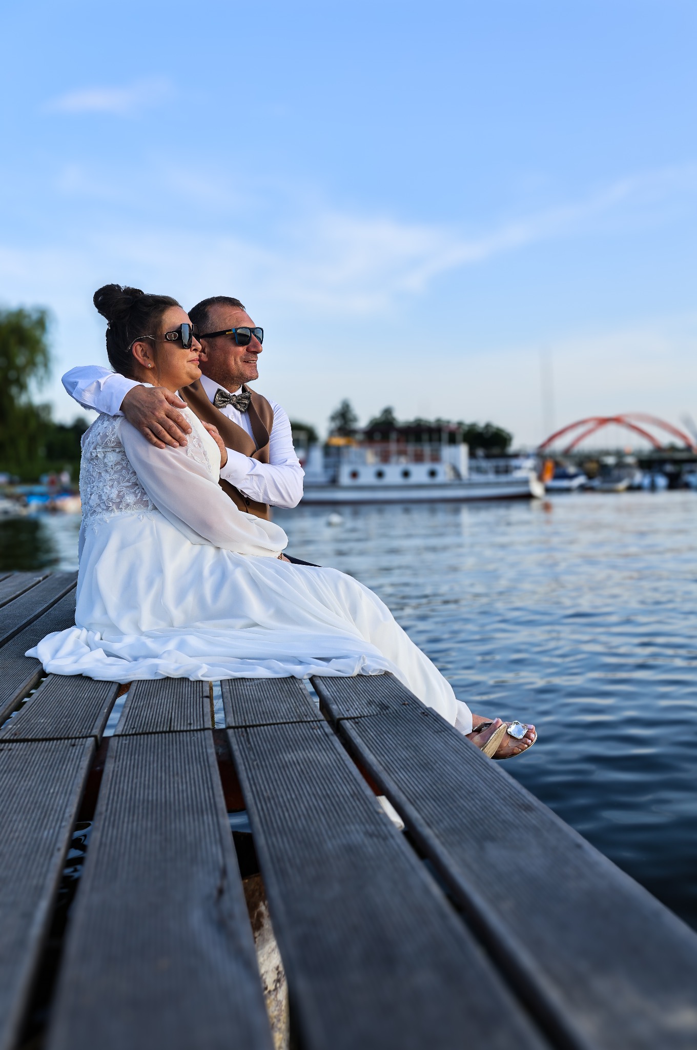 Para młoda w okularach przeciwsłonecznych siedzi na pomoście nad wodą, w tle widoczny most i łodzie. Romantyczna sesja ślubna nad jeziorem.