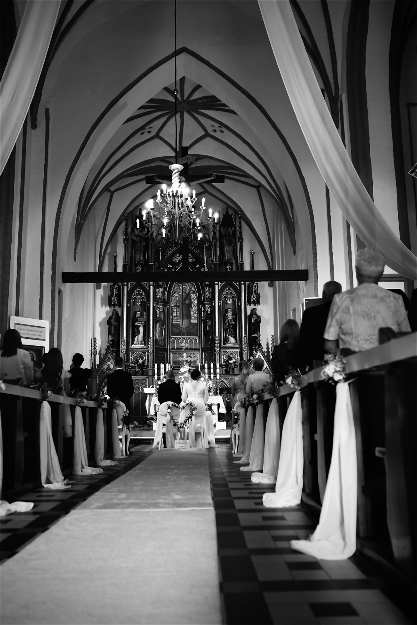 Czarno-białe zdjęcie z uroczystości ślubnej w kościele. Para młoda klęczy przed ołtarzem, widoczny bogato zdobiony ołtarz i goście siedzący w ławkach.