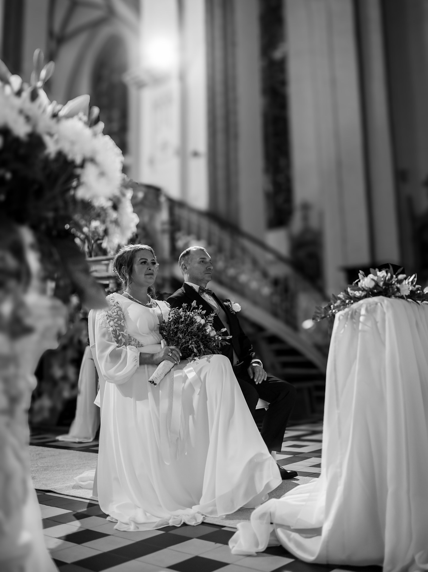 Czarno-białe zdjęcie pary młodej siedzącej w kościele podczas ceremonii ślubnej. Panna młoda trzyma bukiet, a w tle widoczne schody i dekoracje kwiatowe.