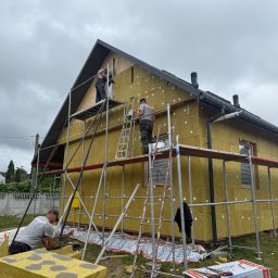 Rem-Bud Nowak - Ocieplanie domu wełną mineralną na rusztowaniu. Widoczne płyty izolacyjne, pracownicy i narzędzia. Dom z ciemnym dachem i oknami w trakcie prac wykończeniowych.