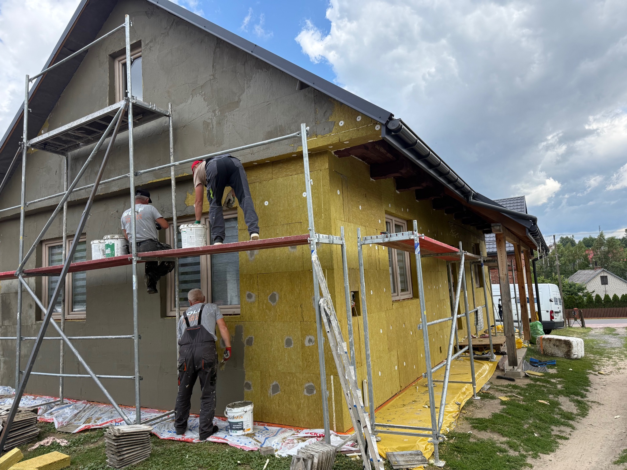Ocieplanie i szpachlowanie elewacji domu jednorodzinnego. Prace na rusztowaniu, widoczna warstwa izolacji i nakładany tynk. Fachowcy w trakcie pracy.