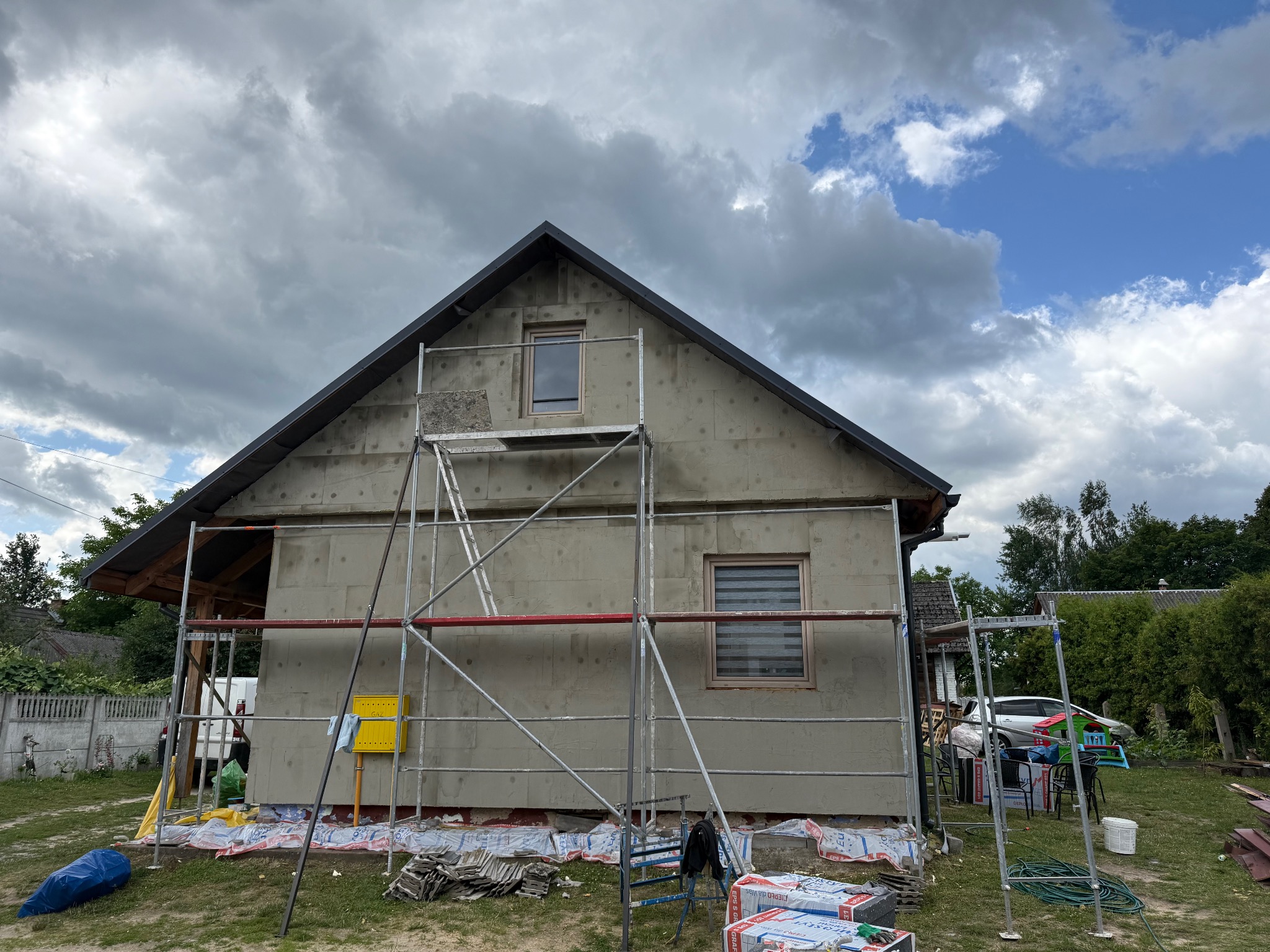 Dom w trakcie ocieplania styropianem, widoczne rusztowania i materiały budowlane. Prace wykończeniowe elewacji w toku, pochmurne niebo w tle.