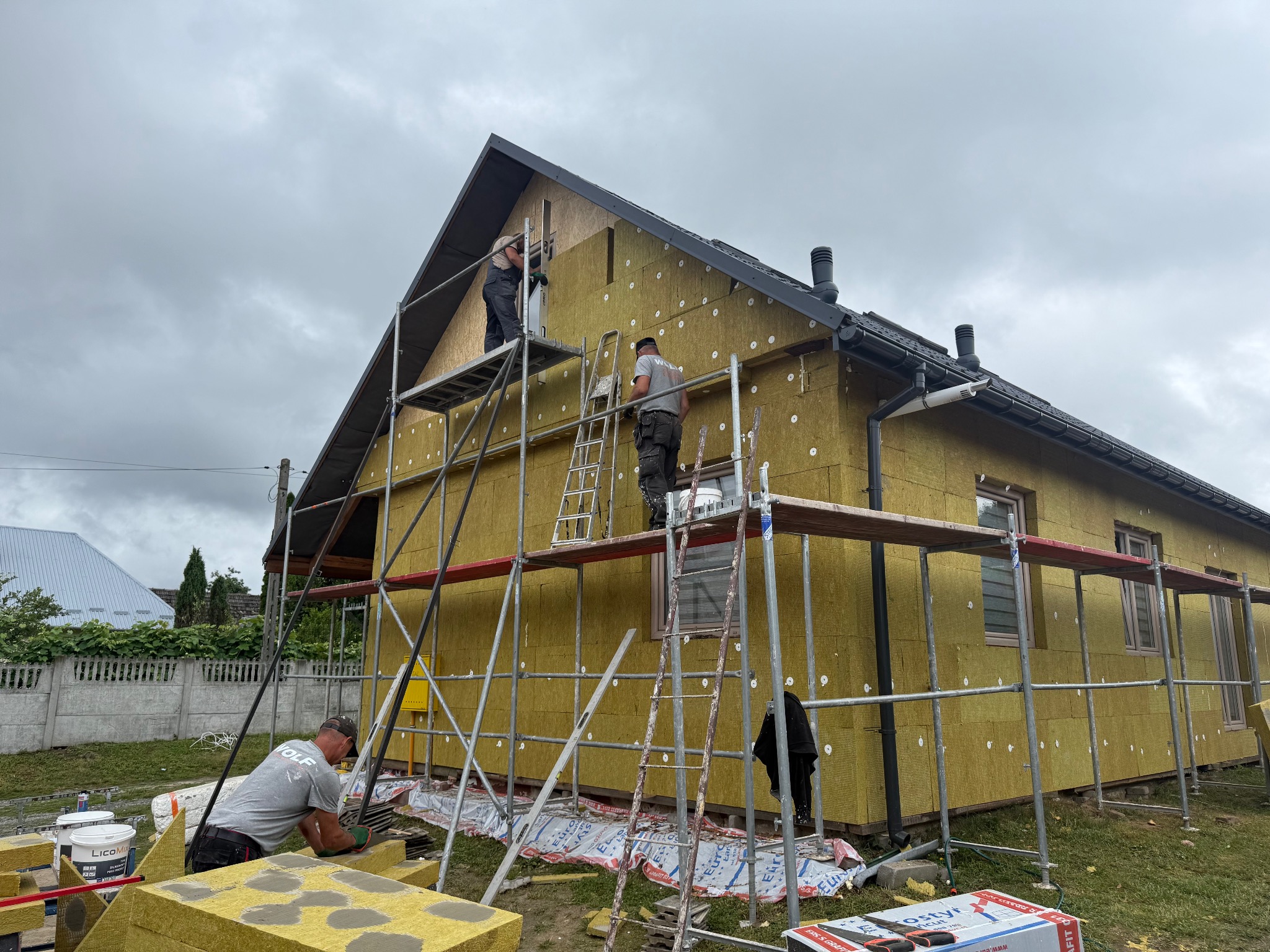 Ocieplanie domu wełną mineralną na rusztowaniu. Widoczne płyty izolacyjne, pracownicy i narzędzia. Dom z ciemnym dachem i oknami w trakcie prac wykończeniowych.