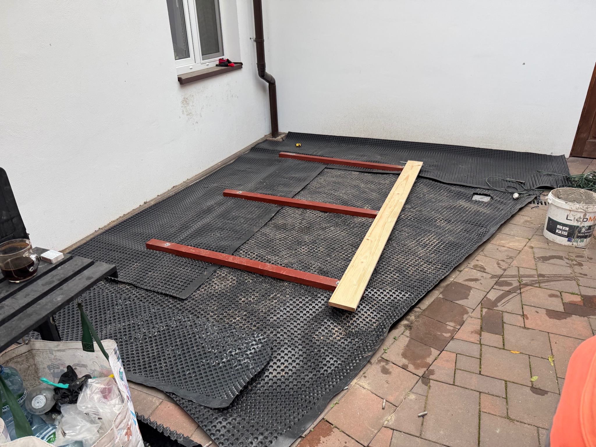 Przygotowanie podłoża: czarna folia drenażowa z wypustkami i drewniane legary na patio. Widoczny fragment ściany budynku i akcesoria budowlane.