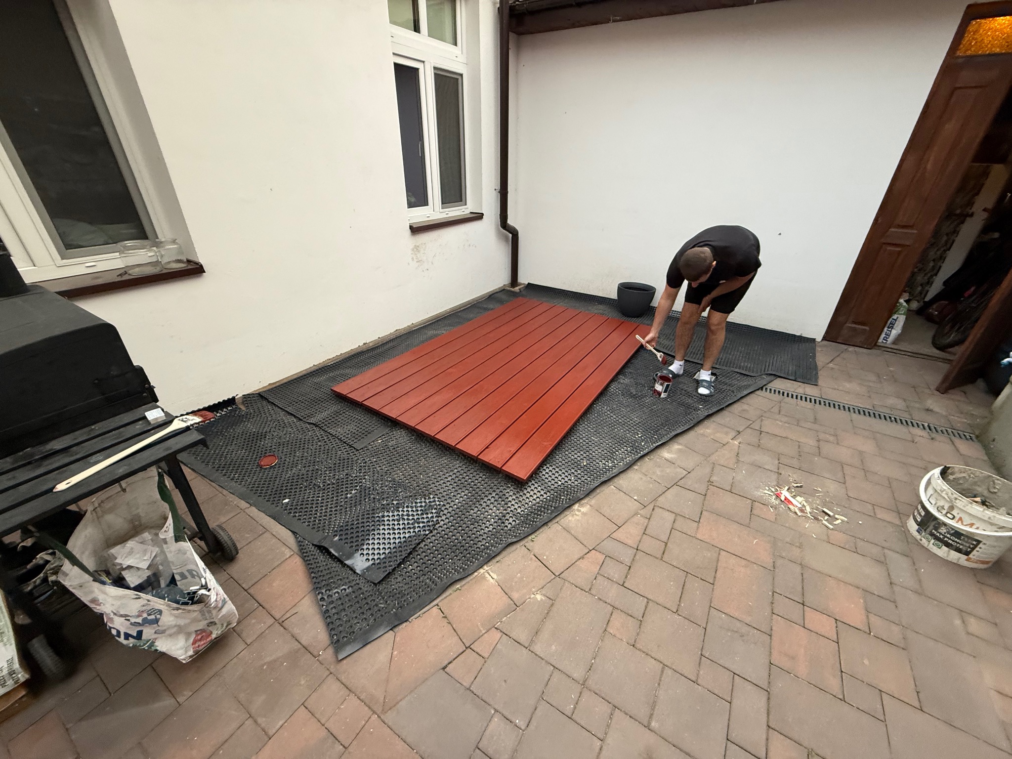 Mężczyzna maluje drewnianą konstrukcję na zewnątrz, widoczne narzędzia malarskie i wiadro z farbą. Praca w toku, ceglana podłoga na zewnątrz.