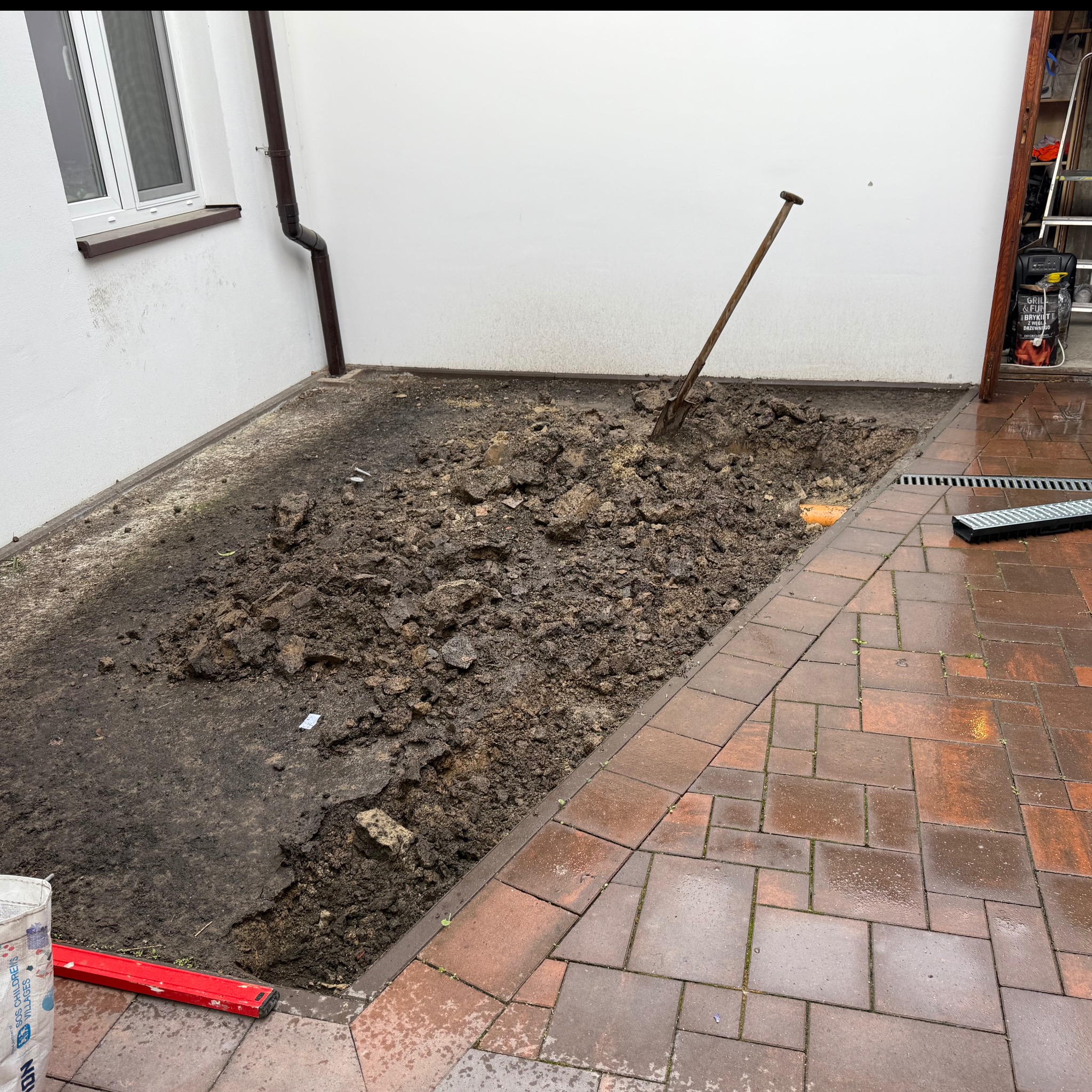 Wykopana ziemia w prostokątnym obszarze obok budynku z ceglaną kostką, łopata wbita w ziemię, widoczny poziomica i worek z zaprawą. Etap przygotowawczy prac ziemnych.
