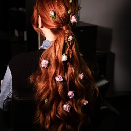 SI SALON SPÓŁKA Z OGRANICZONĄ ODPOWIEDZIALNOŚCIĄ - Rudowłosa kobieta z tyłu, z wplecionymi różowymi różami w warkocz, siedząca na czarnym skórzanym fotelu. Fryzura artystyczna w ciemnym otoczeniu.