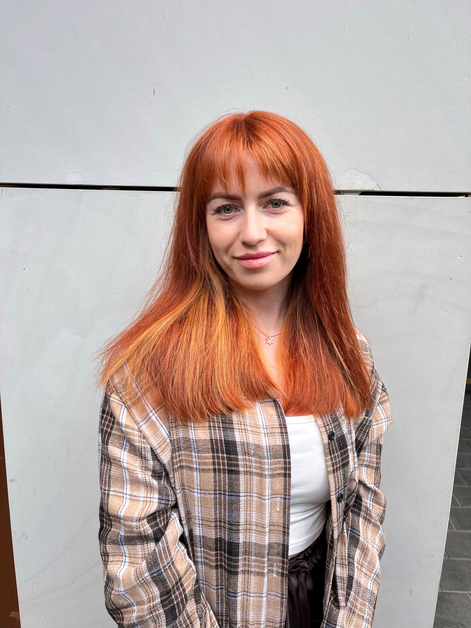 Uśmiechnięta kobieta z długimi, rudymi włosami i grzywką, ubrana w koszulę w kratę, pozuje na tle szarej ściany. Włosy mają jaśniejsze pasemka.