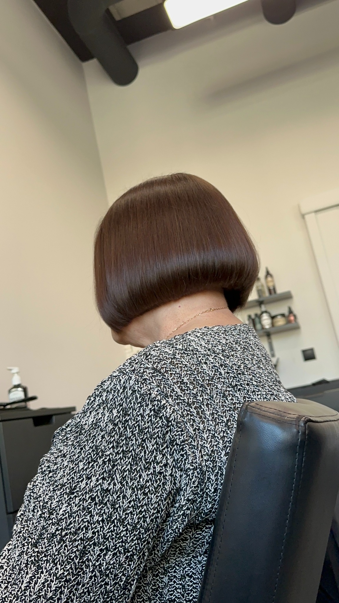 Gładkie, brązowe bob cięcie na kobiecie w melanżowym swetrze, widok z tyłu, złoty łańcuszek na szyi, półka z kosmetykami w tle.