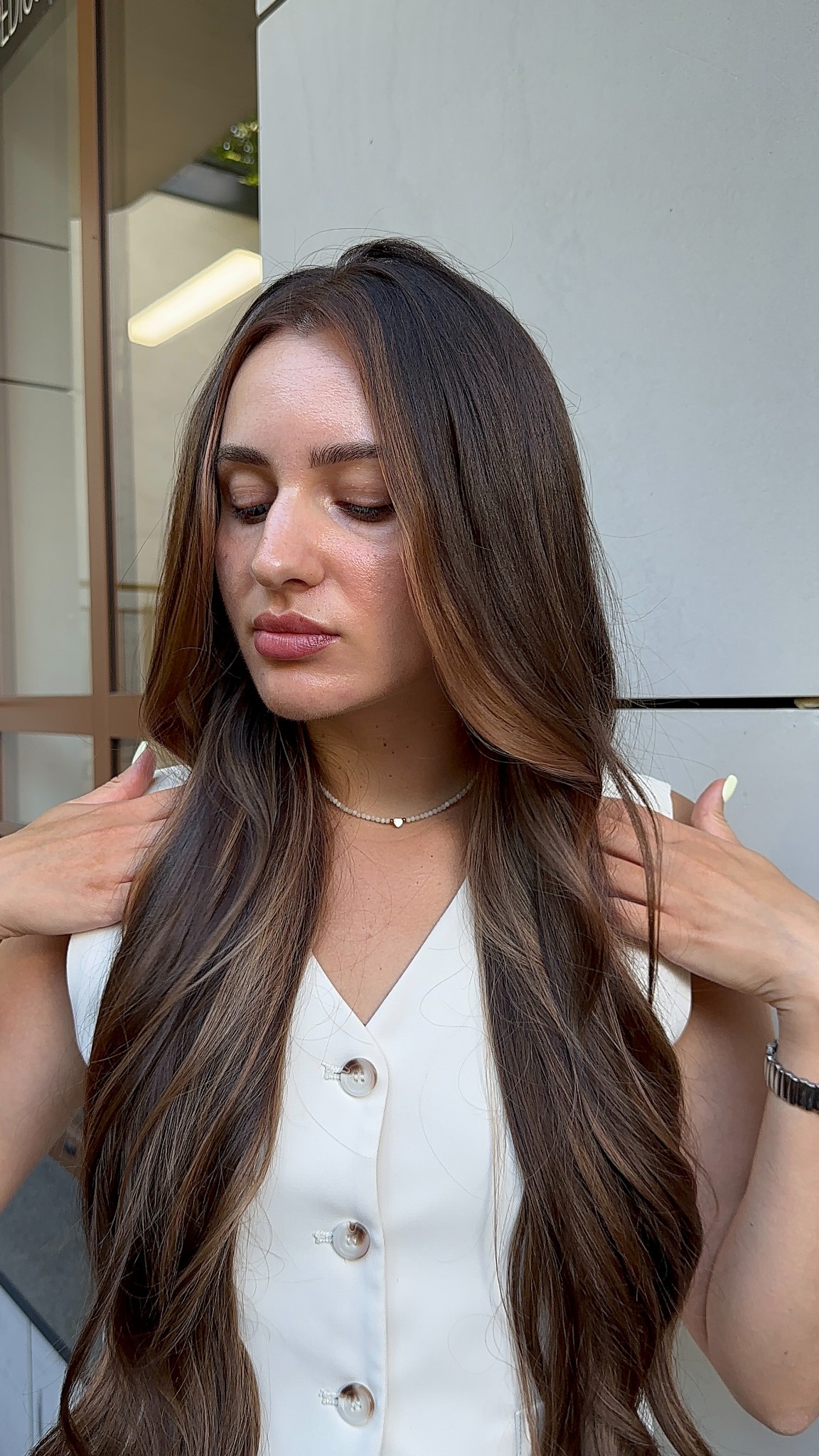 Kobieta z długimi, brązowymi włosami z refleksami, w białej bluzce, pozuje z rękami na ramionach. Widoczne delikatne fale i ombre na włosach.