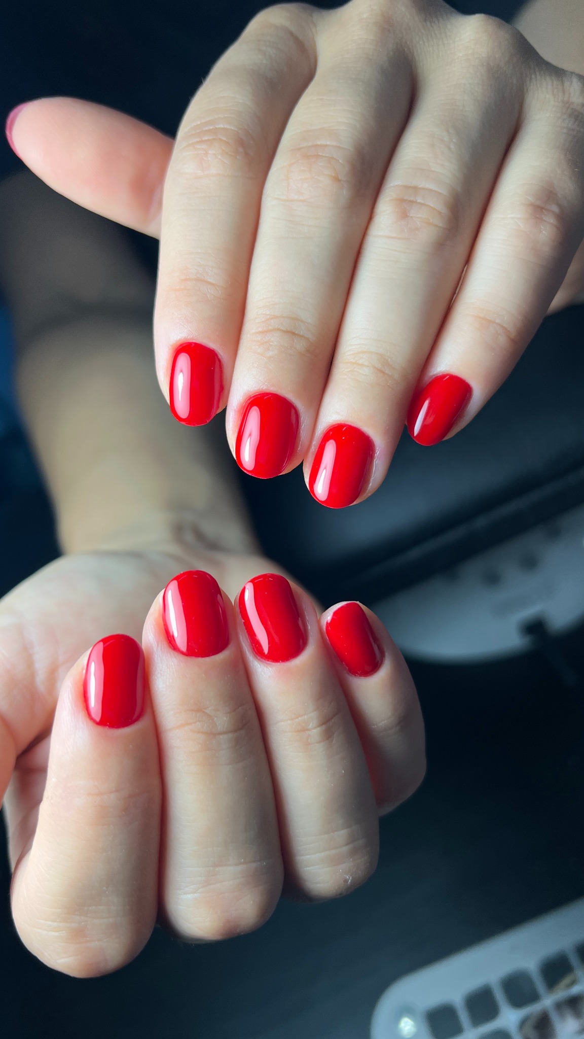 Dłonie z czerwonym, błyszczącym manicure hybrydowym na krótkich, owalnych paznokciach, w zbliżeniu, z widocznym oświetleniem lampy UV.