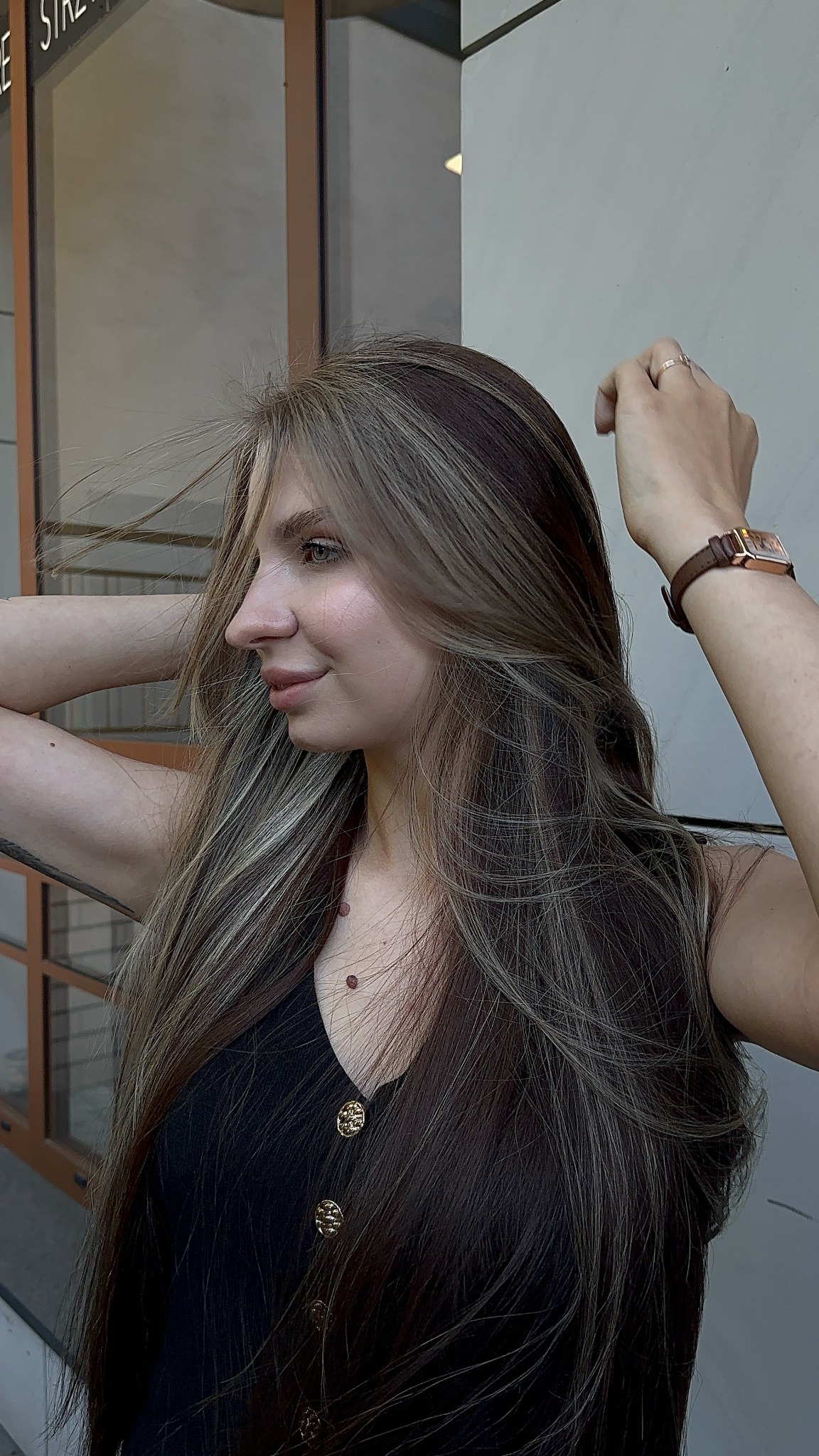 Profil kobiety z długimi, brązowymi włosami z blond pasemkami. Widoczny zegarek na ręce i czarna bluzka z ozdobnymi guzikami. Delikatny makijaż.