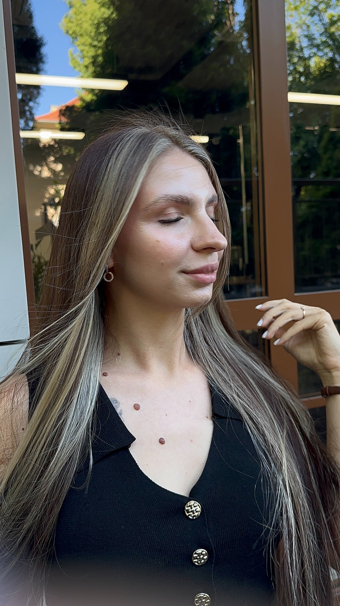 Profil kobiety z długimi, blond włosami z pasemkami, zamkniętymi oczami i delikatnym uśmiechem, w czarnym ubraniu z ozdobnymi guzikami. Widoczne kolczyki i pierścionek.