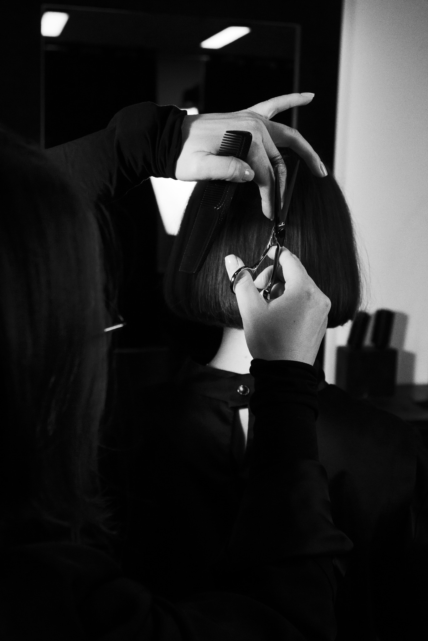 Strzyżenie włosów na krótko w czerni i bieli. W kadrze dłonie fryzjera z grzebieniem i nożyczkami, oraz fragment włosów klientki. W tle widać akcesoria.