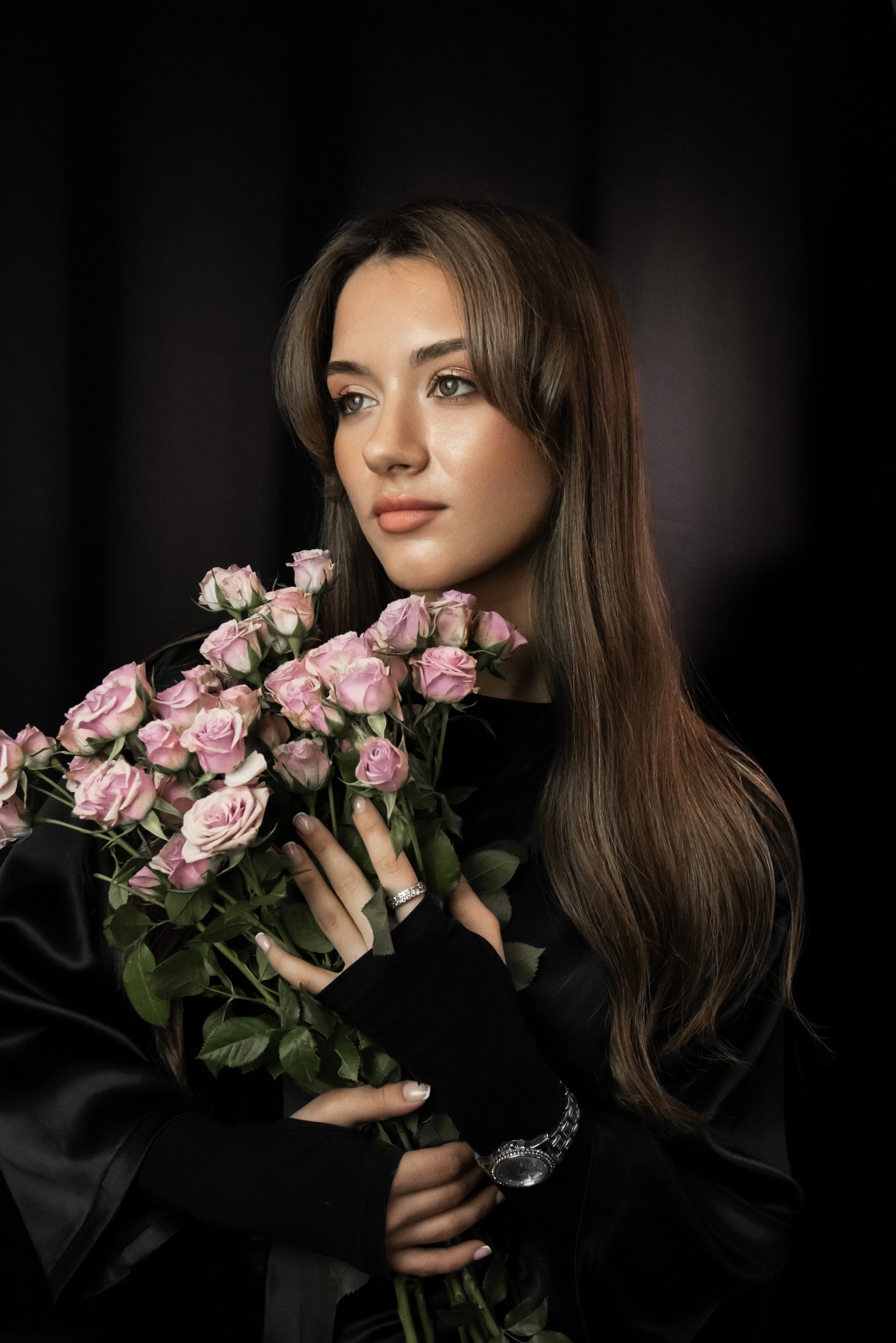 Portret kobiety z długimi, brązowymi włosami, trzymającej bukiet różowych róż na czarnym tle. Subtelny makijaż i manicure dopełniają stylizację.
