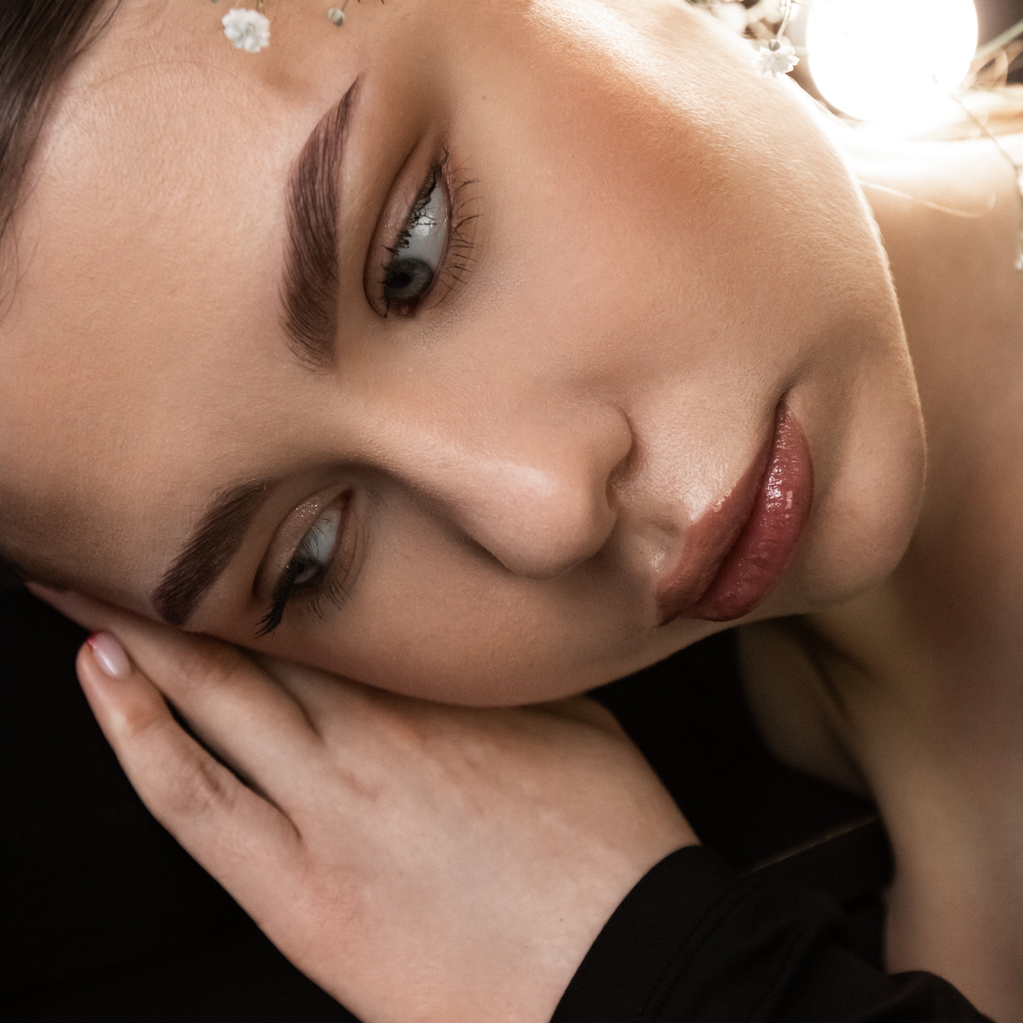 Portret kobiety z delikatnym makijażem, akcentem na brwi i ustach, ozdobiona drobnymi kwiatami we włosach. Ujęcie z bliska, skupione na twarzy.