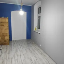 Pokój po malowaniu: błękitna ściana z białym łukiem, szare ściany, jasna podłoga imitująca drewno, okno i wisząca lampa. Widoczny piec kaflowy.