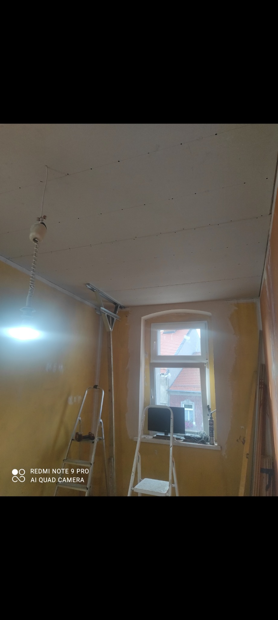 Montaż płyt gipsowo-kartonowych na suficie w pokoju z żółtymi ścianami. Widoczne profile, drabiny i okno. Prace wykończeniowe w trakcie.