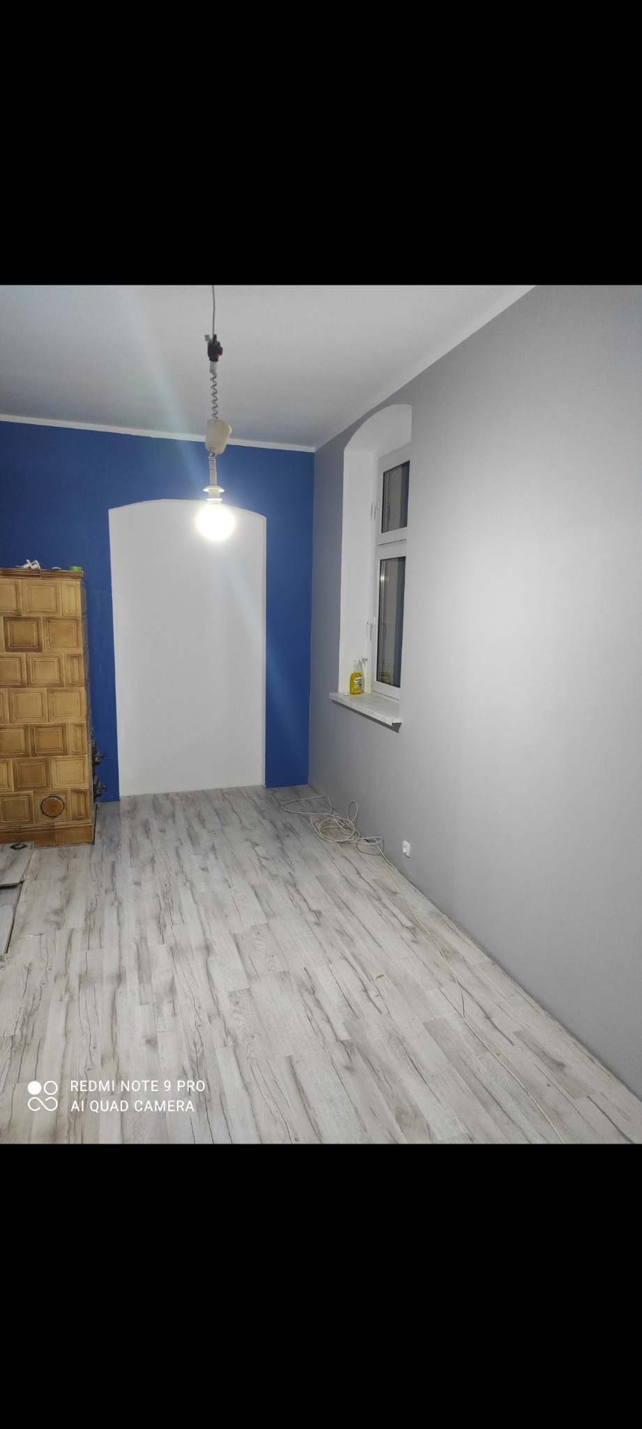 Pokój po malowaniu: błękitna ściana z białym łukiem, szare ściany, jasna podłoga imitująca drewno, okno i wisząca lampa. Widoczny piec kaflowy.