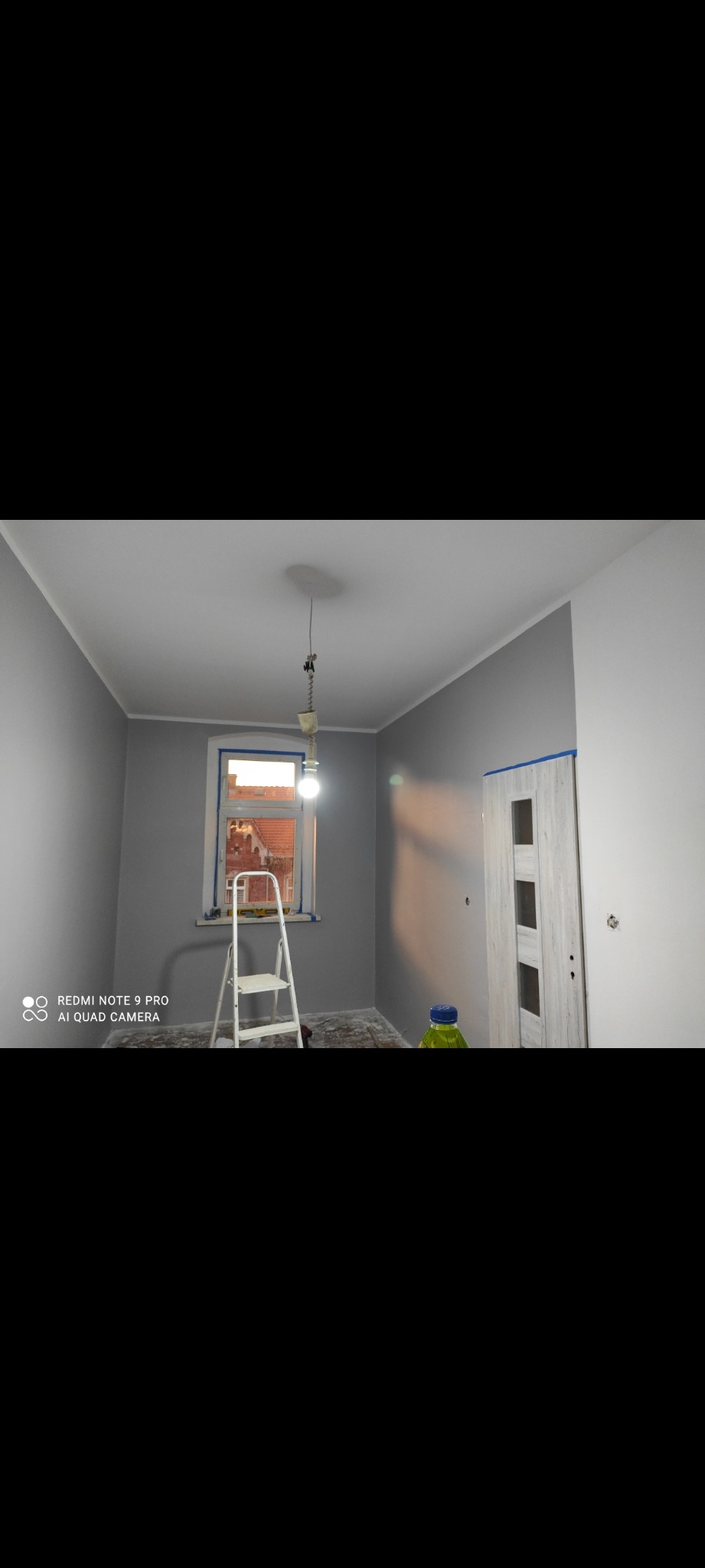 Pokój po malowaniu na szaro, z drabiną przy oknie i butelką farby. Widoczne taśmy malarskie na ościeżnicach okna i drzwi. Całość w neutralnej tonacji.