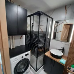 Nowoczesna łazienka z kabiną prysznicową, umywalką nablatową i pralką pod blatem. Marmurowe płytki na ścianach, czarne meble i elementy wykończenia.