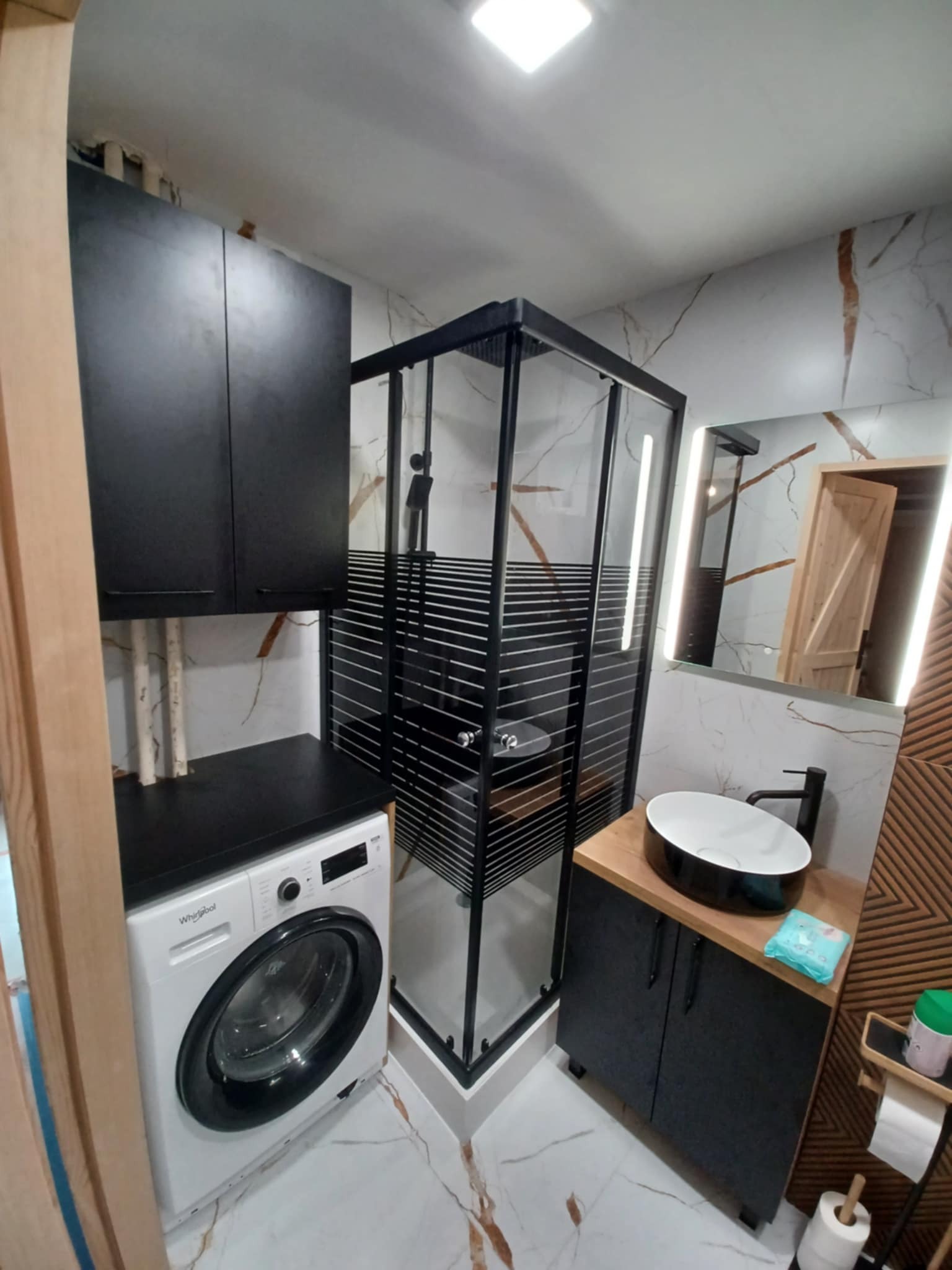 Nowoczesna łazienka z kabiną prysznicową, umywalką nablatową i pralką pod blatem. Marmurowe płytki na ścianach, czarne meble i elementy wykończenia.