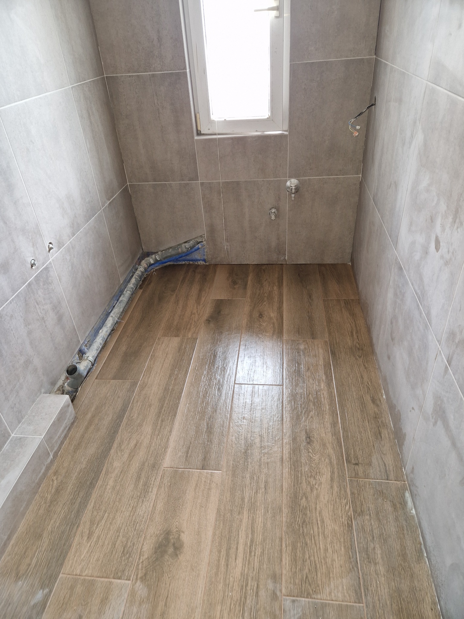 Wykończona podłoga z płytek imitujących drewno w szarej łazience z oknem. Widoczne fragmenty instalacji wodnej w ścianie.