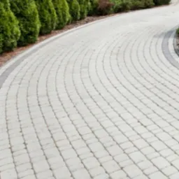 Zakrzywiony podjazd z jasnoszarej kostki brukowej, obrzeża z ciemniejszej kostki. Wzdłuż podjazdu rząd wysokich, gęstych tuj.