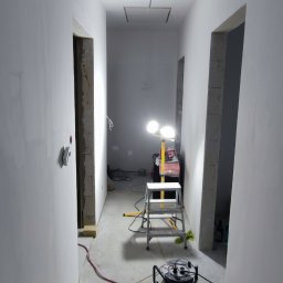 Renofin - Korytarz w trakcie prac wykończeniowych: świeżo wyszpachlowane ściany, widoczne kable, drabina i lampa budowlana. Fragment podłogi z ułożonym parkietem.