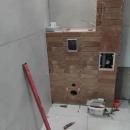Wnętrze łazienki w trakcie wykańczania. Ułożone płytki ścienne i podłogowe. Widoczne narzędzia glazurnicze, poziomica. Fragment instalacji wodnej w ścianie.