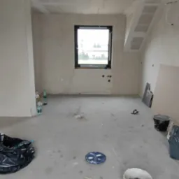 Surowe wnętrze z oknem, ściany i sufit wykończone płytami gipsowo-kartonowymi, przygotowane do dalszych prac wykończeniowych. Widoczne narzędzia i materiały budowlane.