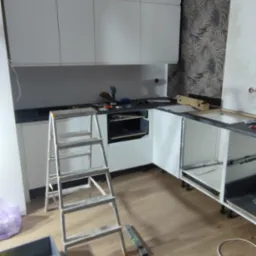 Montaż białej, nowoczesnej kuchni. Widoczne szafki, drabina, narzędzia i fragment tapety w liście na ścianie. Etap prac: instalacja mebli kuchennych.