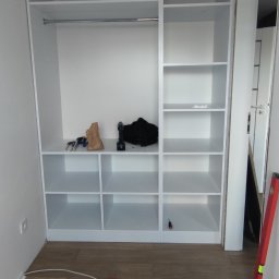 Renofin - Biała, wnękowa szafa z półkami i drążkiem na ubrania w trakcie montażu, na podłodze leżą narzędzia i poziomica.