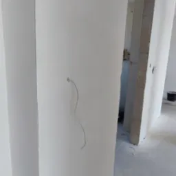Biała ściana po szpachlowaniu z wystającym przewodem elektrycznym i widocznym wejściem do innego pomieszczenia w trakcie wykończenia.