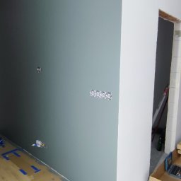 Renofin - Wnętrze w trakcie wykańczania: ściana pomalowana na szaro-zielono z gniazdkami elektrycznymi, podłoga z panelami. Widoczne narzędzia i kartony po materiałach.