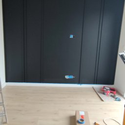 Renofin - Ściana z czarnymi panelami dekoracyjnymi w trakcie montażu, widoczne gniazdka elektryczne, narzędzia i lampa sufitowa w minimalistycznym stylu.