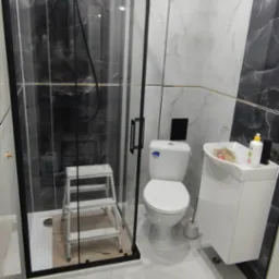 Wykończona łazienka z kabiną prysznicową typu walk-in z czarnymi ramami, białą armaturą i marmurowymi płytkami. Widoczna drabina malarska w kabinie.