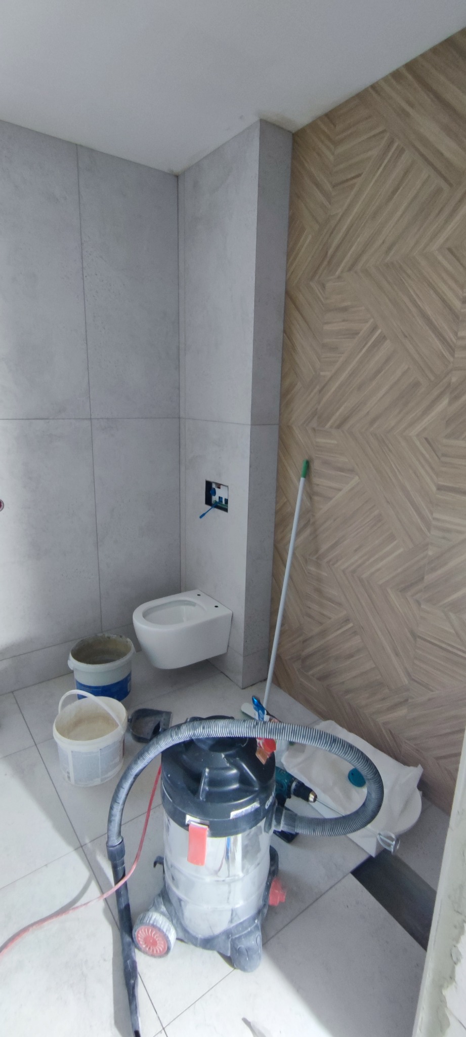 Remont łazienki: częściowo wykończona, nowoczesna łazienka z płytkami imitującymi beton i drewno, wisząca toaleta i odkurzacz przemysłowy na podłodze.