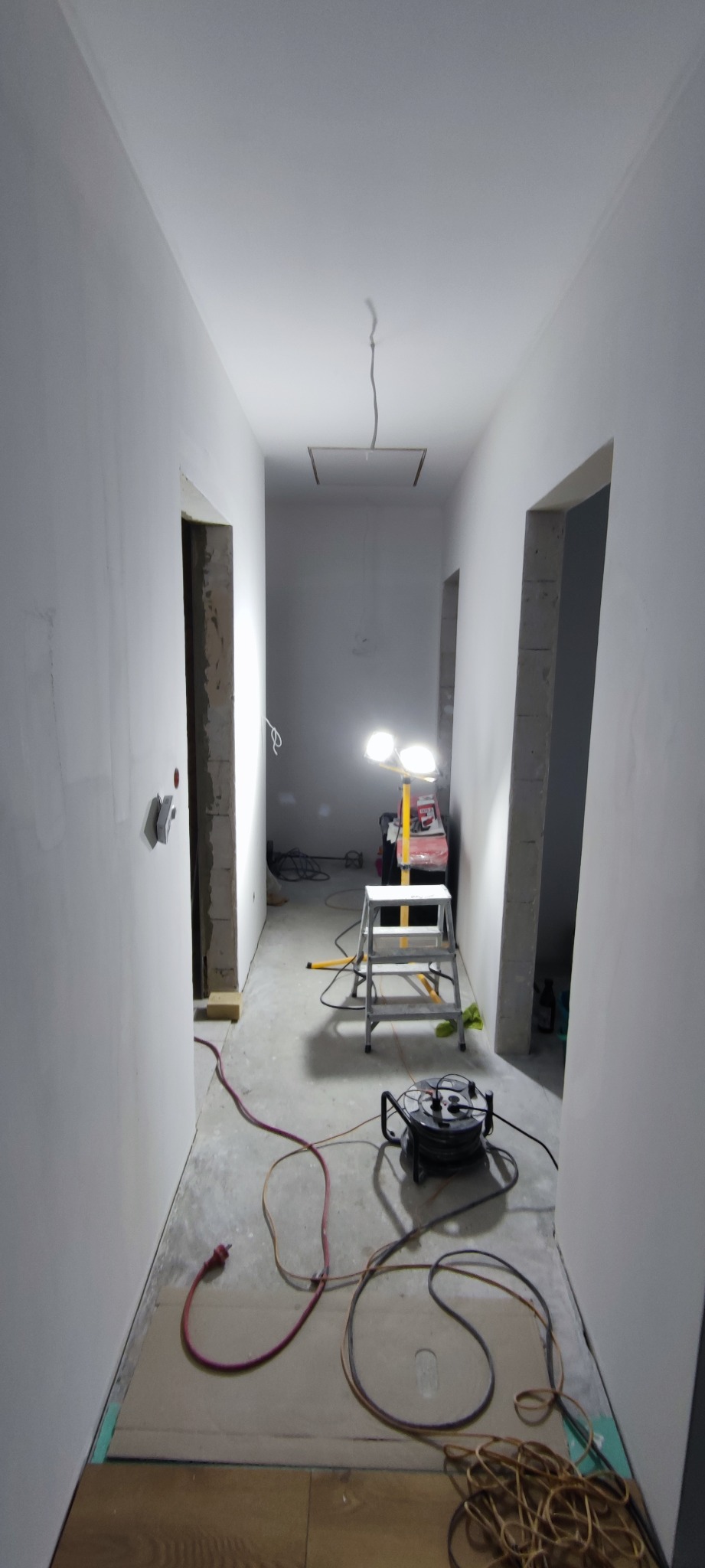 Korytarz w trakcie prac wykończeniowych: świeżo wyszpachlowane ściany, widoczne kable, drabina i lampa budowlana. Fragment podłogi z ułożonym parkietem.