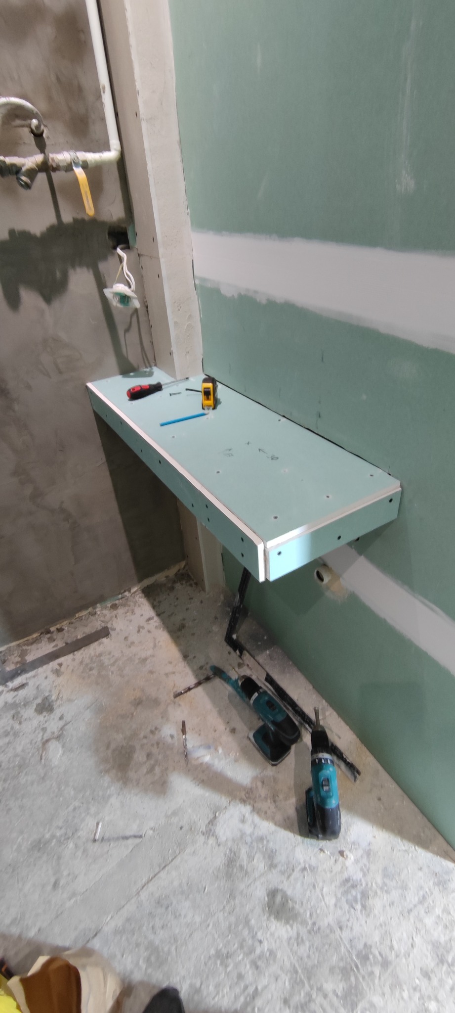 Montaż półki z płyty karton-gips w surowym wnętrzu. Widoczne narzędzia, ściany w trakcie wykańczania, rury. Półka przymocowana do ściany z płyt g-k.