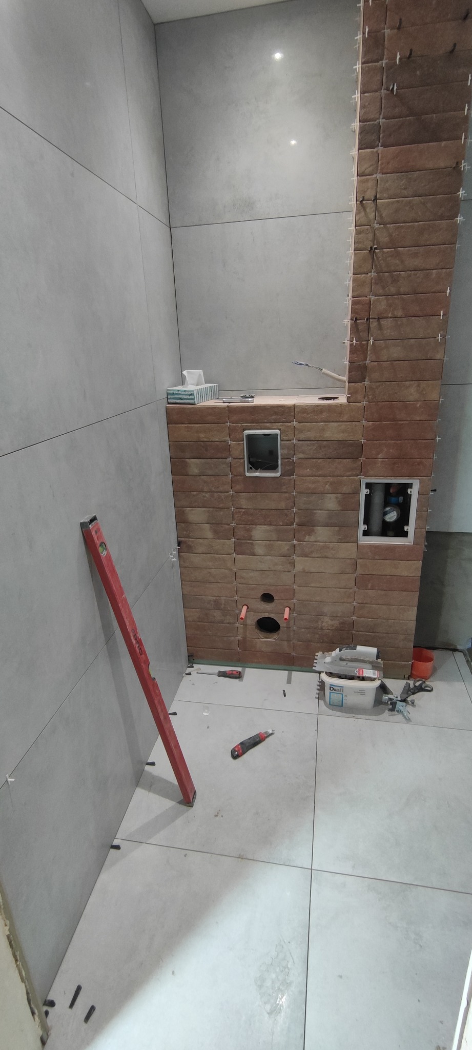 Wnętrze łazienki w trakcie wykańczania. Ułożone płytki ścienne i podłogowe. Widoczne narzędzia glazurnicze, poziomica. Fragment instalacji wodnej w ścianie.