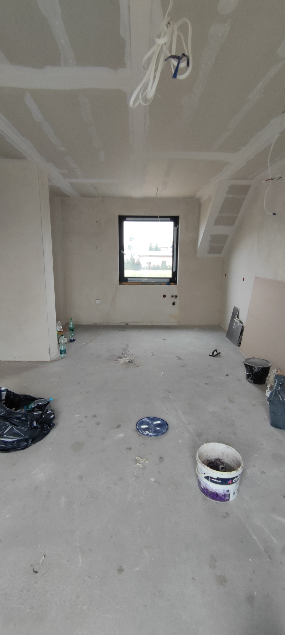 Surowe wnętrze z oknem, ściany i sufit wykończone płytami gipsowo-kartonowymi, przygotowane do dalszych prac wykończeniowych. Widoczne narzędzia i materiały budowlane.