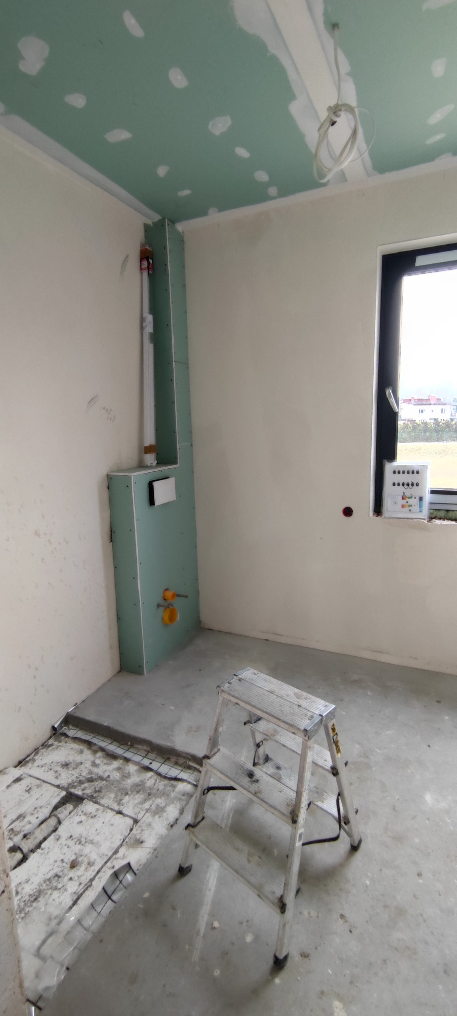 Łazienka w trakcie prac: zabudowa z płyty KG zakrywająca instalacje, fragment ogrzewania podłogowego, drabina. Widoczne elementy instalacji sanitarnej.
