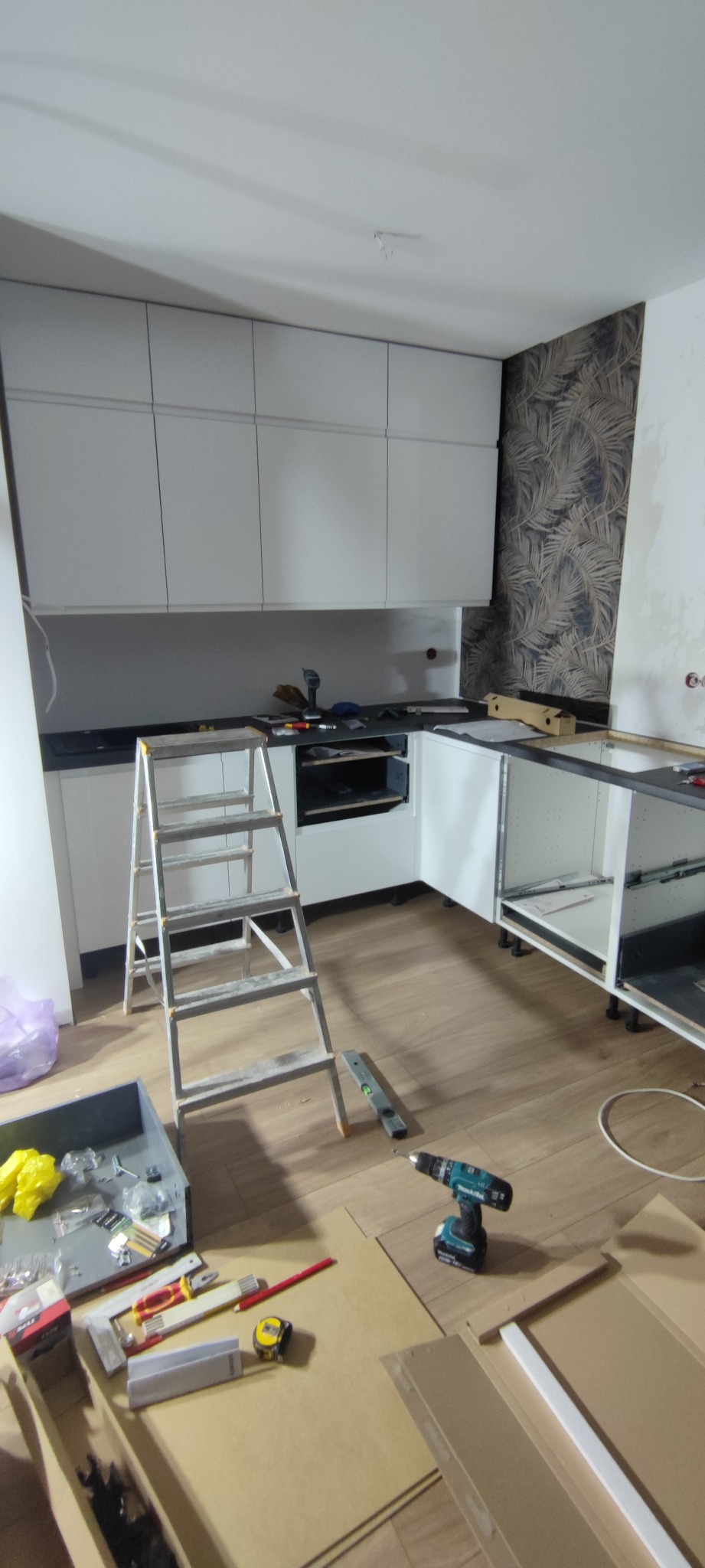 Montaż białej, nowoczesnej kuchni. Widoczne szafki, drabina, narzędzia i fragment tapety w liście na ścianie. Etap prac: instalacja mebli kuchennych.