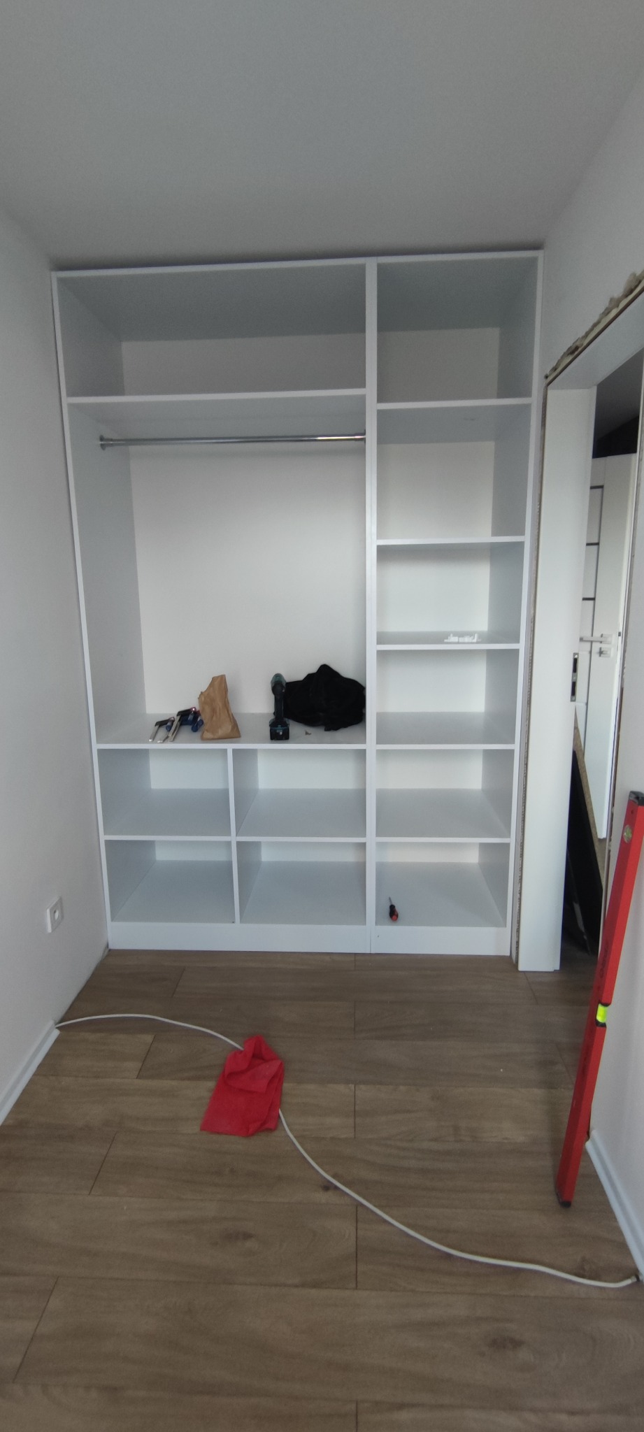 Biała, wnękowa szafa z półkami i drążkiem na ubrania w trakcie montażu, na podłodze leżą narzędzia i poziomica.