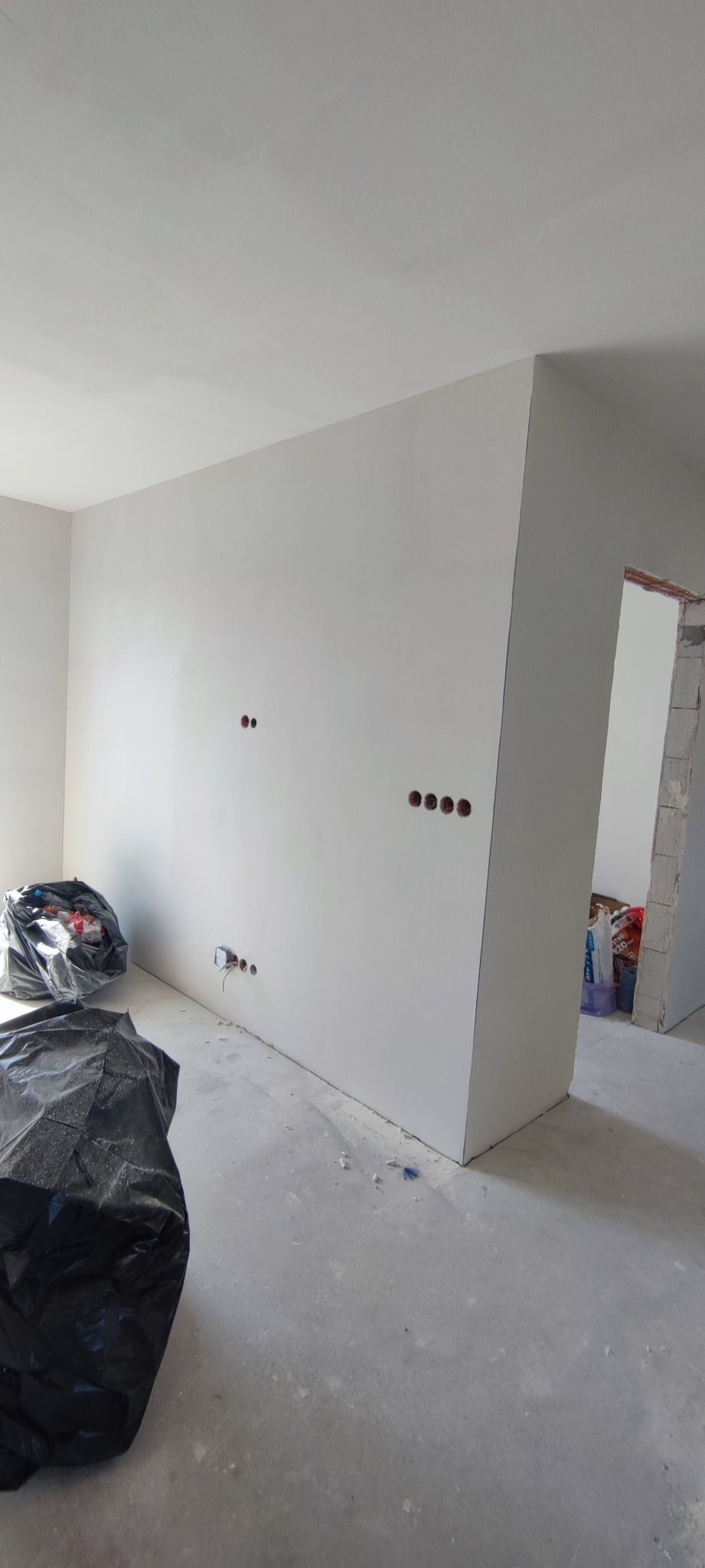 Wnętrze w trakcie szpachlowania: gładkie, białe ściany z widocznymi otworami na gniazdka elektryczne. Na podłodze resztki materiałów budowlanych i worki na śmieci.